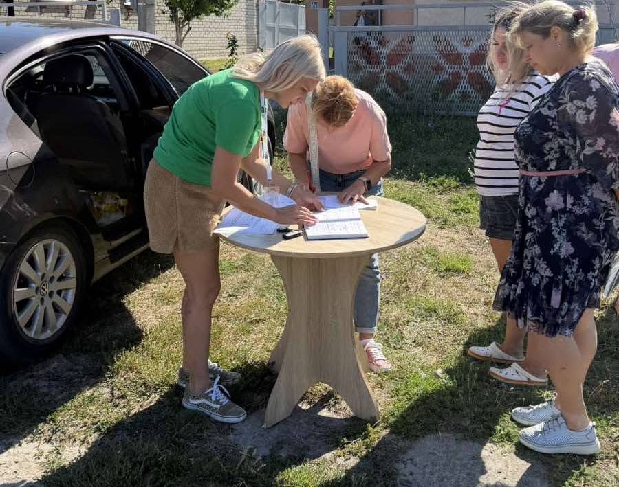 Жінки в черзі заповнюють документи на дерев'яному столі біля автомобіля.