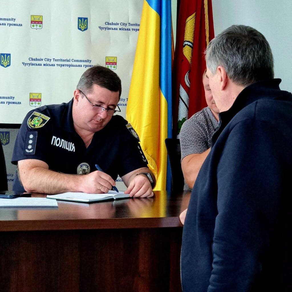 Поліцейський у формі підписує документи за столом на тлі прапорів та банера Чугуївської міської територіальної громади.