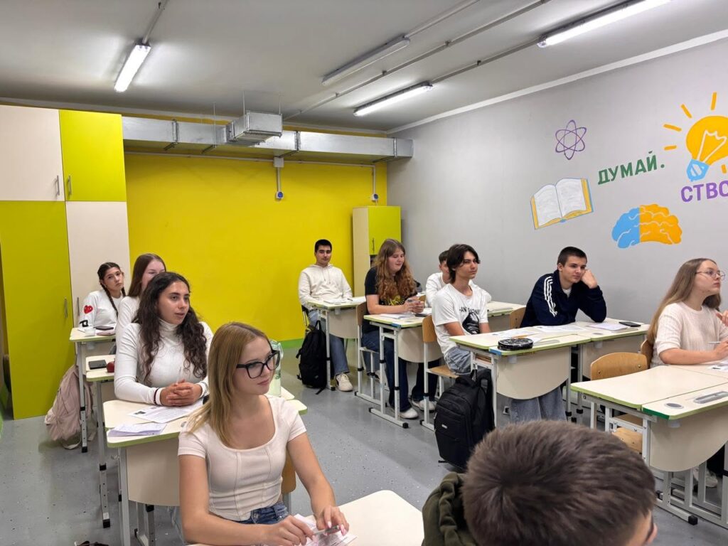 Учні сидять за партами в класі з жовтими стінами та написом «ДУМАЙ. СТВОРЮЙ.» на тлі.