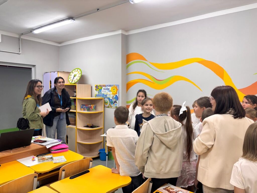 Діти та дорослі спілкуються в класі з жовтими партами та яскравим орнаментом на стіні.