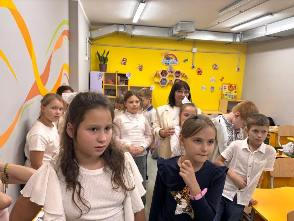 Група дітей та доросла жінка стоять у яскравому класі з жовтими стінами та дитячими прикрасами.