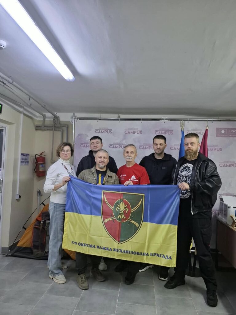 Група людей тримає прапор 129 окремої важкої механізованої бригади на тлі банера з написом CAMPUS.