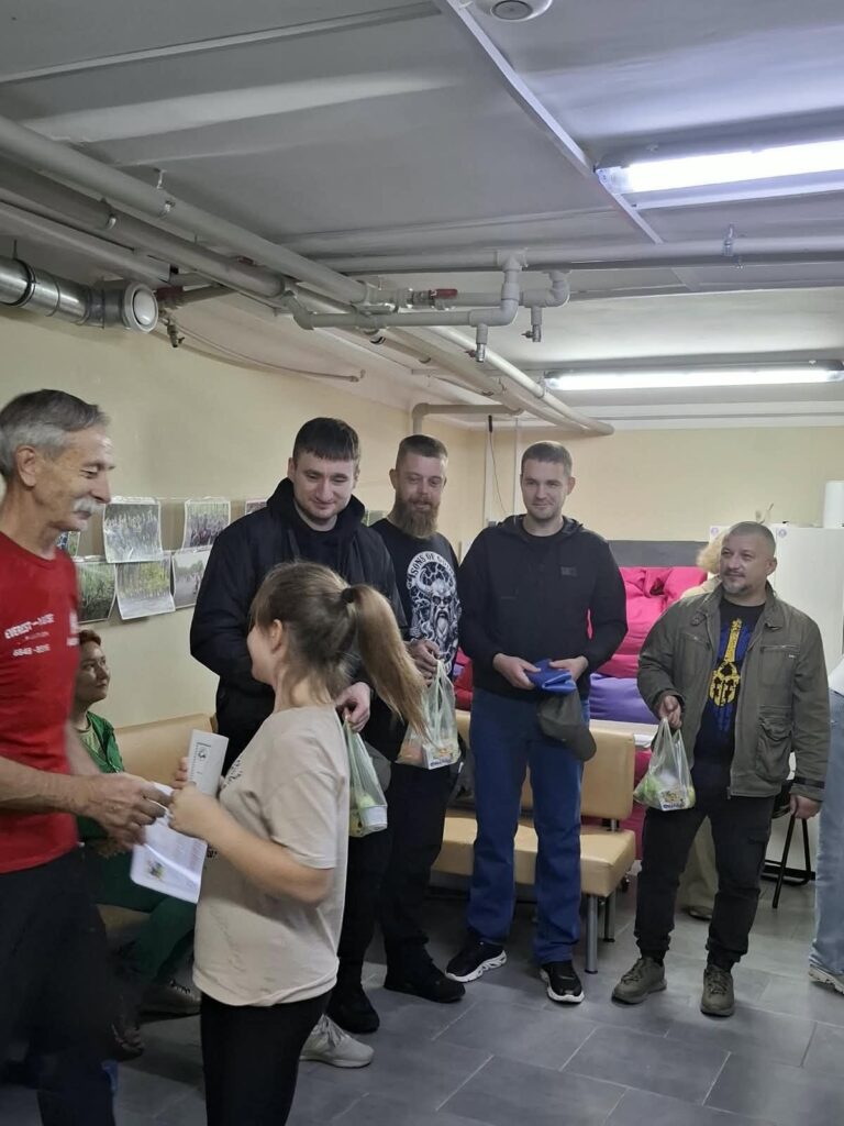 Група людей у приміщенні, де чоловік у червоній футболці видає жінці документи, а інші тримають пакети з продуктами.