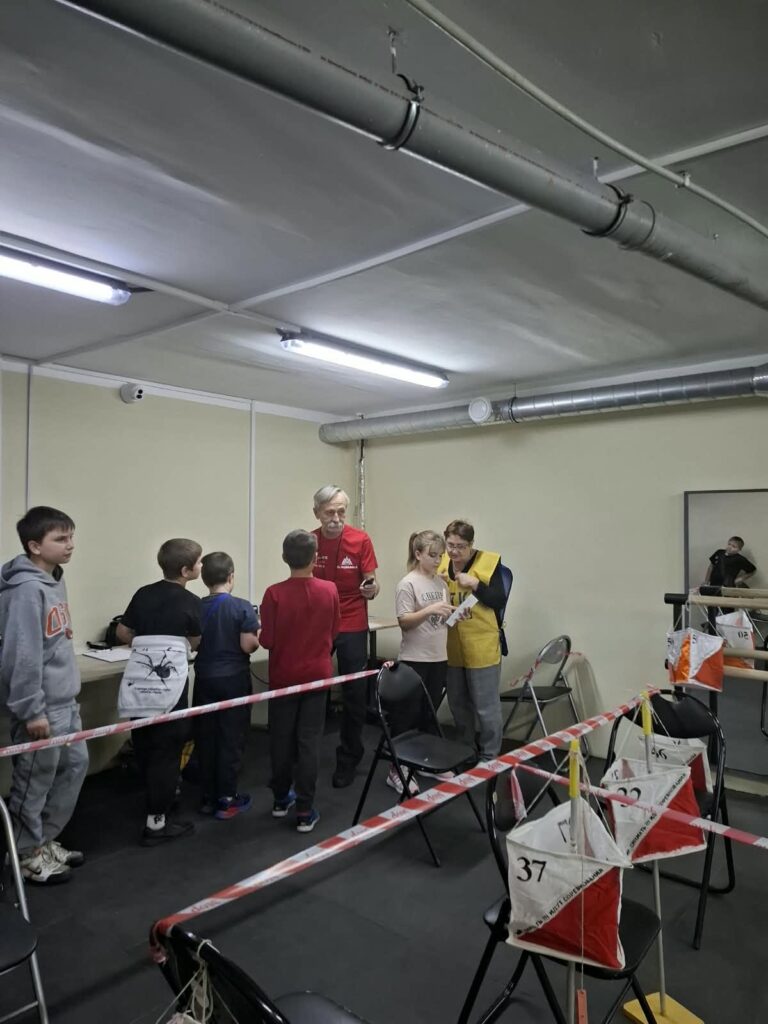Діти та дорослі збирають і запускають паперові дельтаплани з номерами на стільцях у приміщенні.