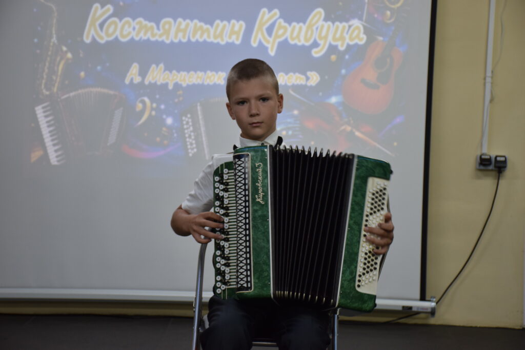Хлопчик Костянтин Кривуца грає на зеленому баяні перед екраном з написом його імені.
