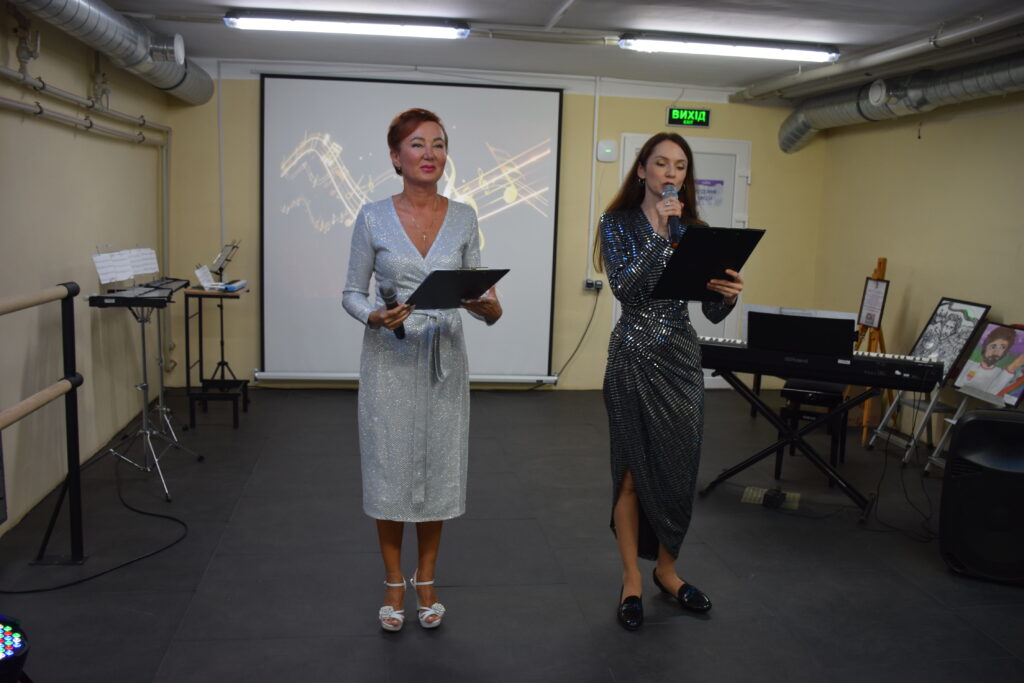 Дві жінки в святкових сукнях стоять на сцені, тримаючи мікрофони та папки, на тлі екрана з музичними нотами.
