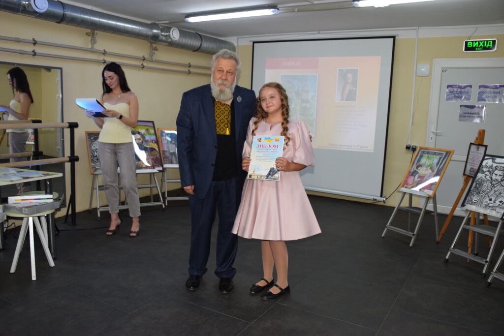` tags. 2. **Analyze the Image:** * **Main Subjects:** An older man with a white beard and a young girl with braided hair. * **Action:** They are standing together, posing for a photo. The girl is holding a document. * **Context/Setting:** It looks like an art exhibition or an award ceremony. There are paintings on easels in the background, a projector screen, and another woman standing nearby reading from a folder. * **Key Details:** * The man is wearing a blue suit jacket and a patterned shirt. * The girl is wearing a pink dress. * The document the girl is holding has the word 