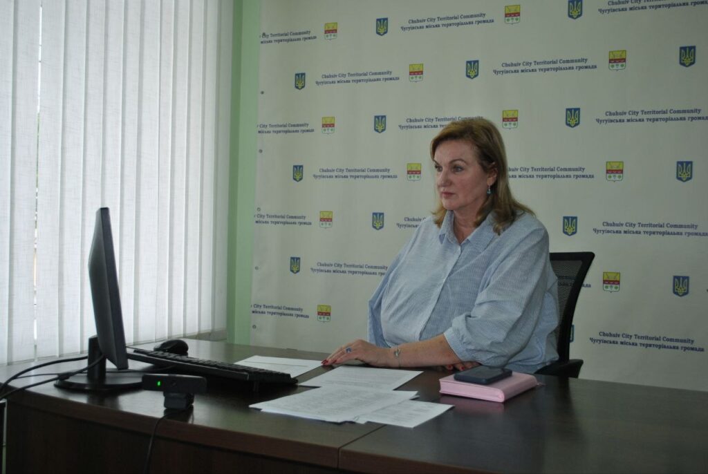 Жінка сидить за столом з документами та комп'ютером на тлі банера з написом 