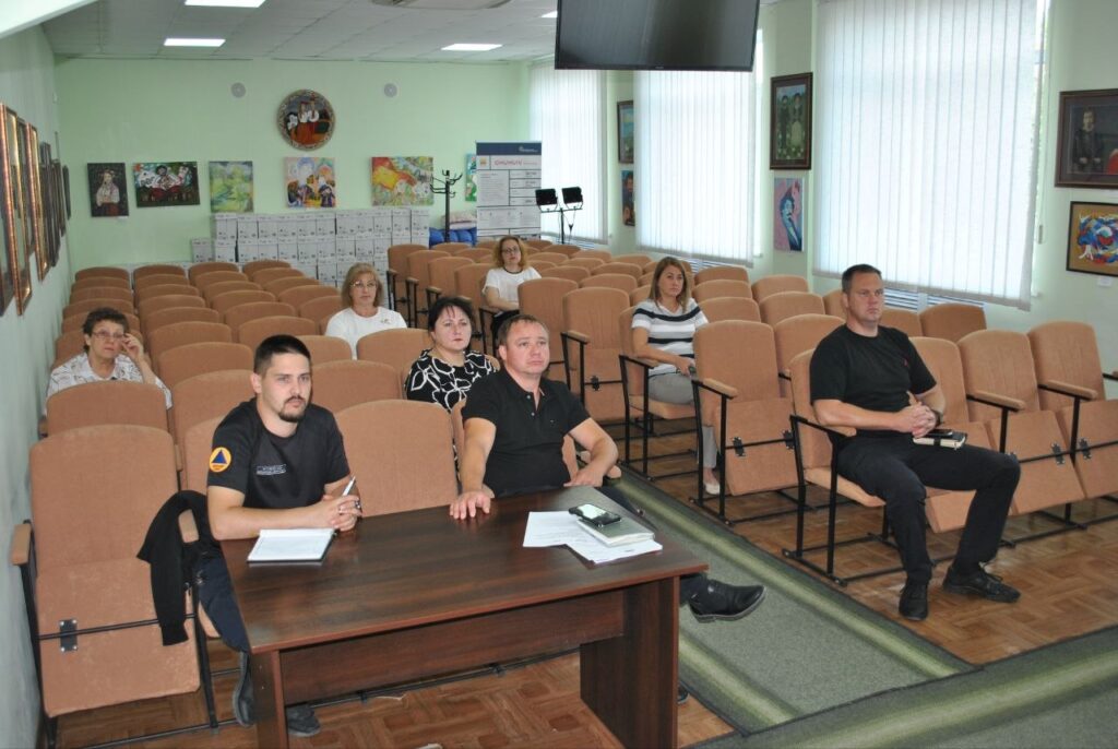 Учасники засідання сидять у залі з рядами крісел, а на передньому плані двоє чоловіків за столом ведуть обговорення.
