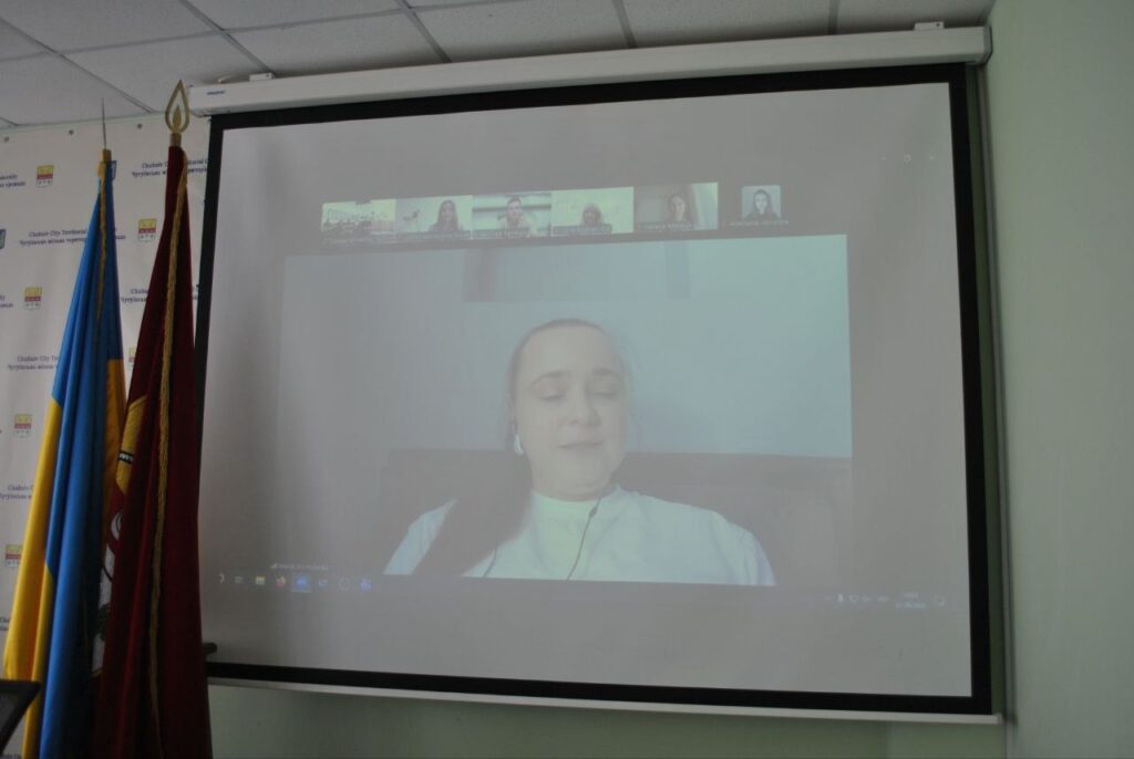Проекційний екран з відеоконференцією, де жінка в білій сорочці з навушниками говорить, а збоку стоять прапори.