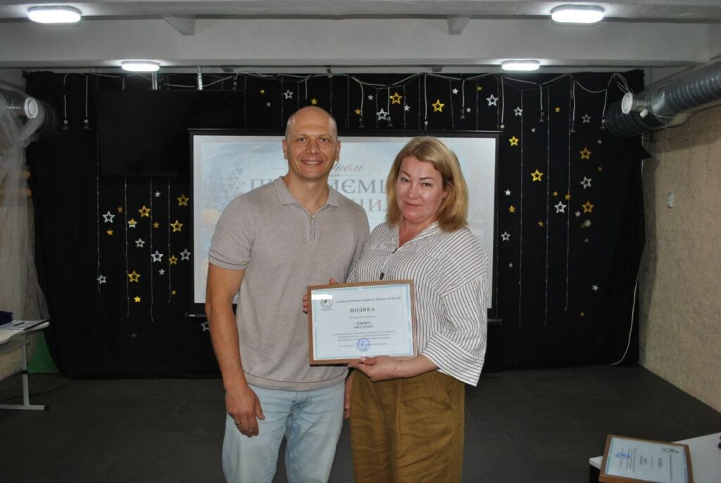 Чоловік і жінка тримають сертифікат із написом «ПОДЯКА» на тлі сцени, прикрашеної світлявими зірками.