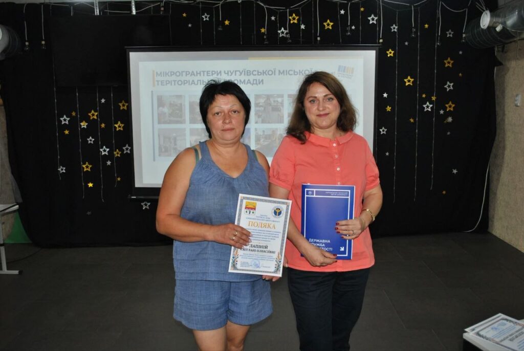Дві жінки тримають нагородні документи «Подяка» та «Державна служба якості» перед банером з написом «Мікрогрантери Чугуївської міської територіальної громади».