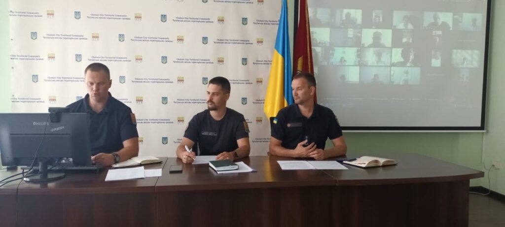 Три чоловіки в формі сидять за столом під час відеоконференції, на екрані за ними видно учасників, а на фоні — прапори та банер з логотипами.