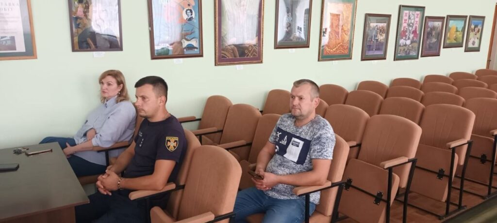 Три людини сидять у залі з рядами порожніх крісел, а на стіні за ними висять рамки з портретами.