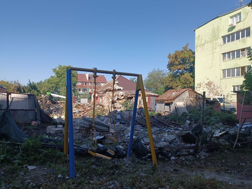Зруйнований дитячий майданчик з пошкодженими гойдалками серед уламків зруйнованих будівель.