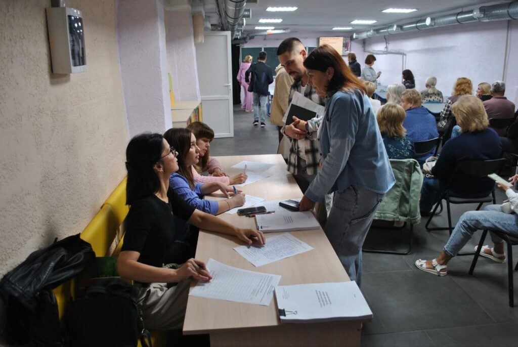 Група людей сидить за столом і заповнює документи, тоді як інші стоять поруч з папками в залі з аудиторією.