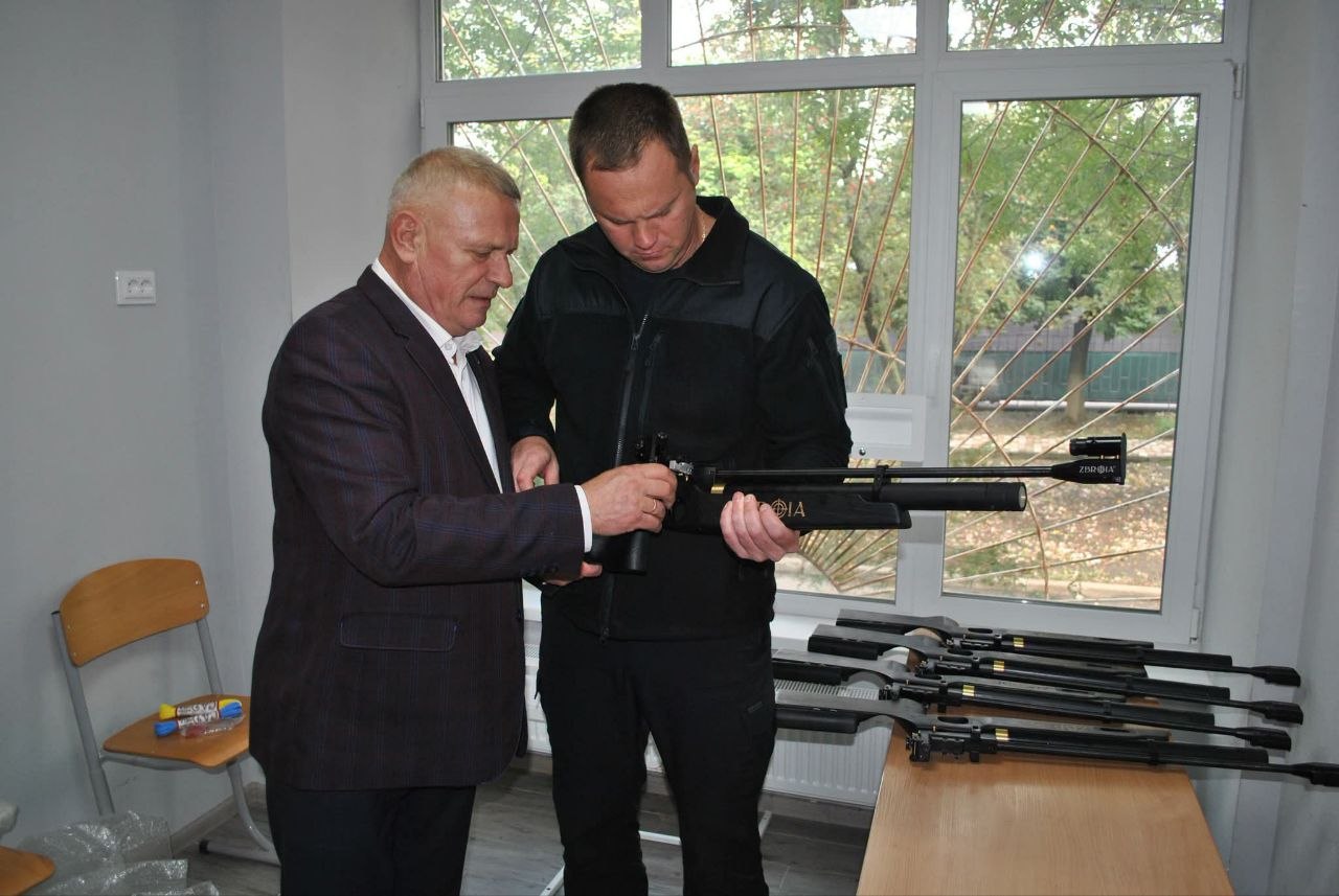 Два чоловіки оглядають і демонструють функціонал чорного гвинтівкового прицілу з написом 