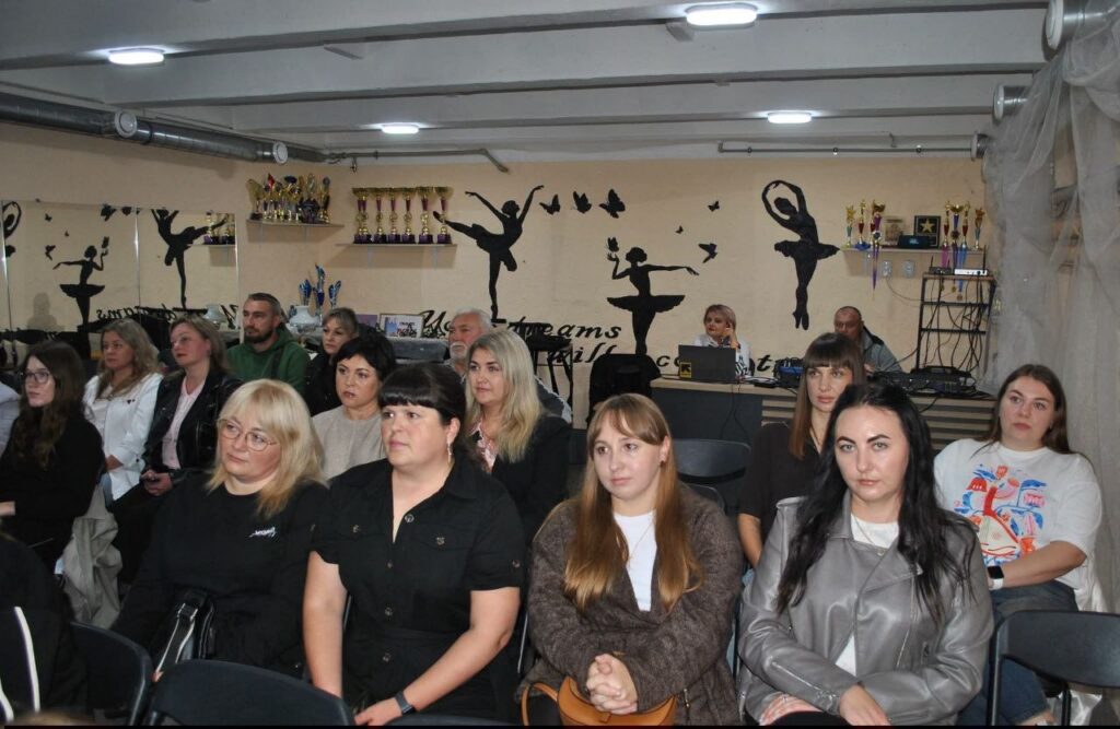 Група людей сидить у залі з силуетами балерин на стіні та поличками з кубками.