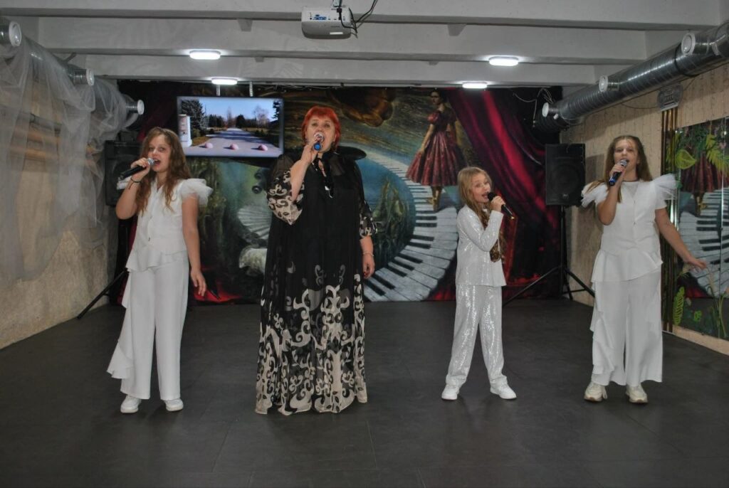 Жінка та три дівчини в білих костюмах співають у мікрофони на сцені.