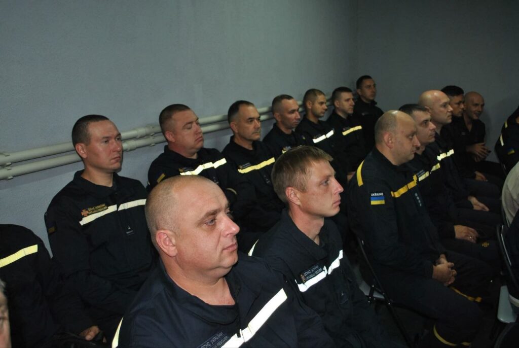 Група рятувальників у чорних формах з жовтими смугами та прапором України сидить рядами та уважно слухає.