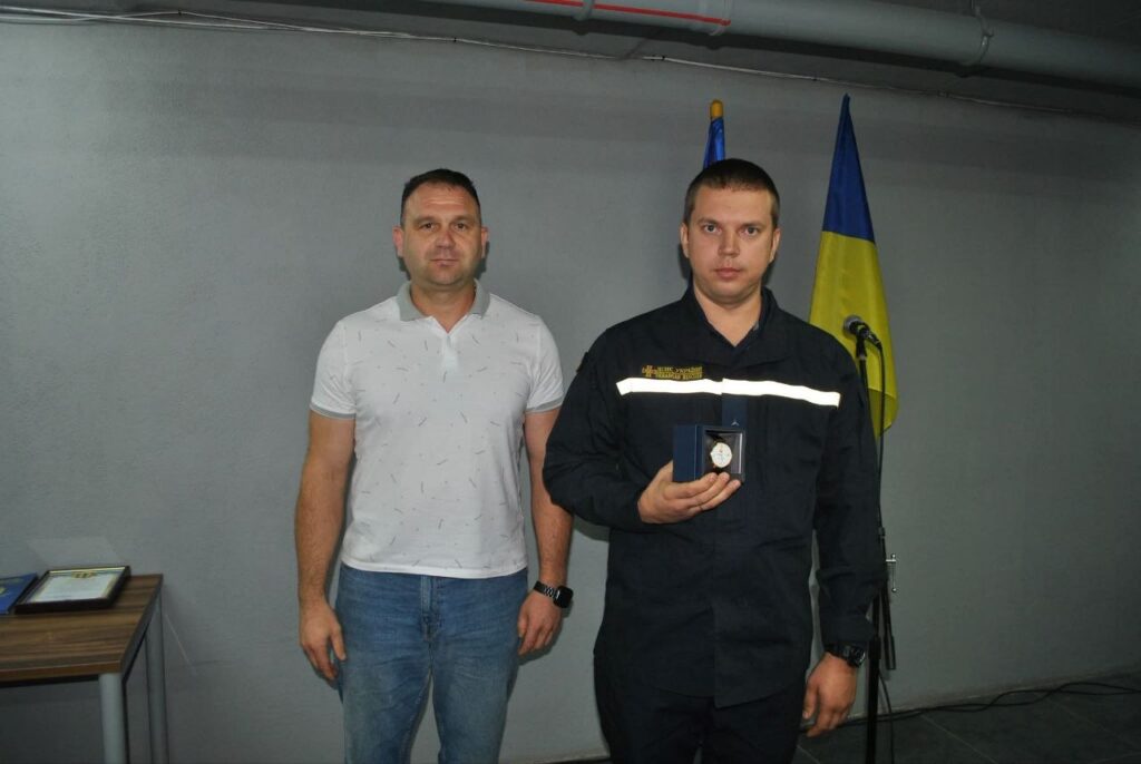 Два чоловіки стоять біля українського прапора, один у формі тримає нагороду в синьому футлярі.