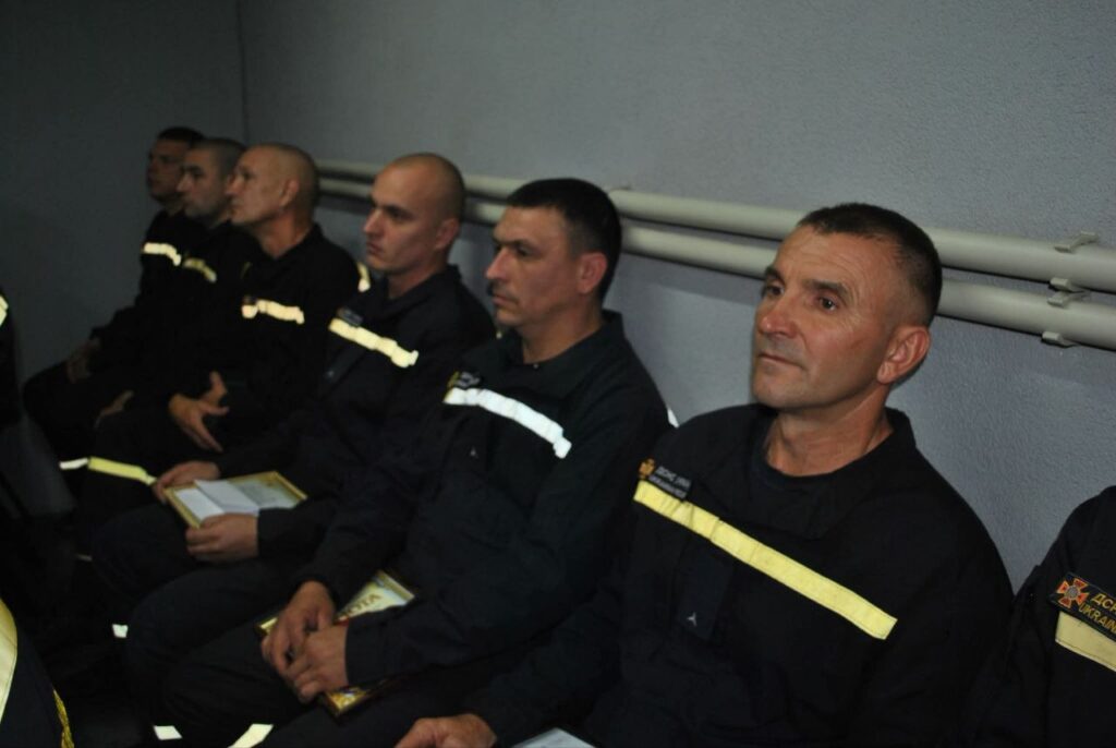 Група рятувальників у чорних формах з жовтими смугами сидить у ряд, слухаючи доповідь.