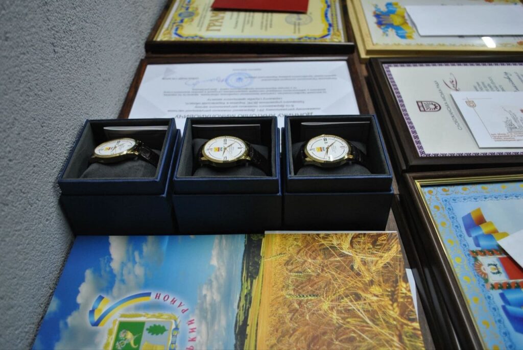 Золоті наручні годинники в чорних коробках, розташовані поруч з рамками з грамотами та фотокартками.