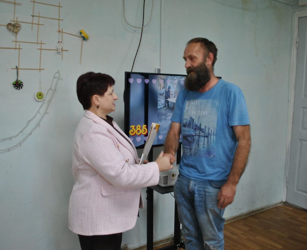 Жінка в рожевому піджаку та чоловік у синій футболці тримають руки над електронною урною для голосування з дисплеєм, на якому видно цифру 385.