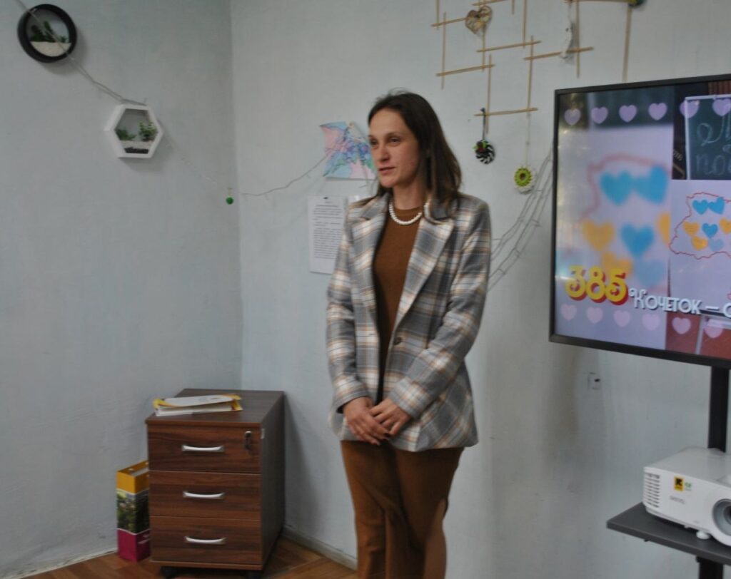 Жінка в клітчастому піджаку стоїть біля екрана з написом «885 ЮЧЕТОВ».