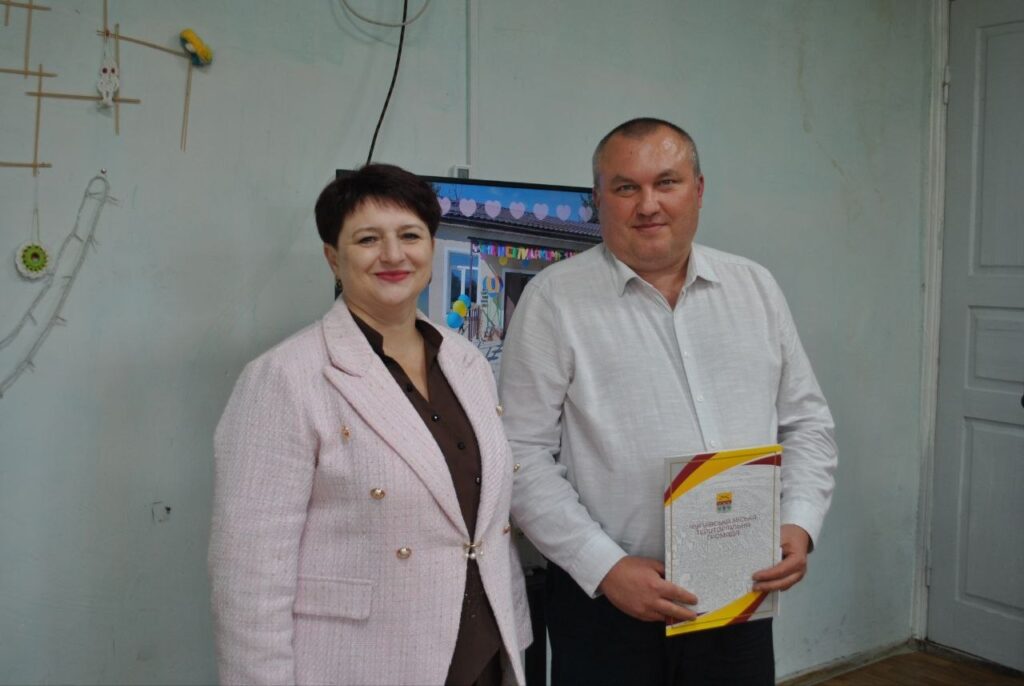 Чоловік у білій сорочці тримає нагороду, стоячи поруч із жінкою в рожевому піджаку.