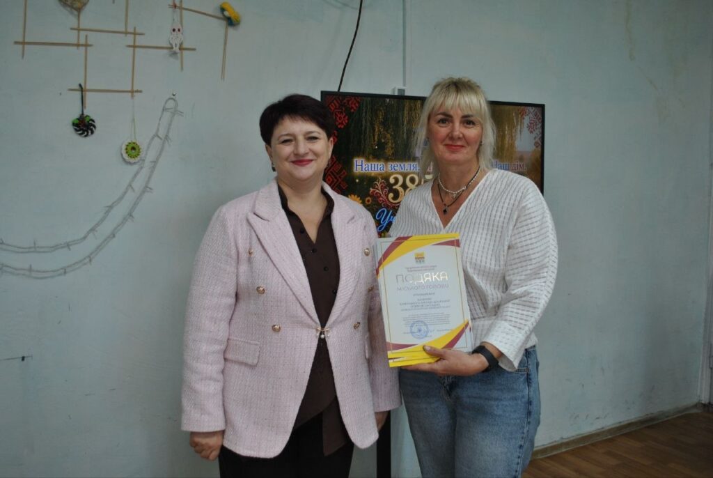Дві жінки тримають нагороду з написом «ПОДЯКА» на тлі стіни з декором.