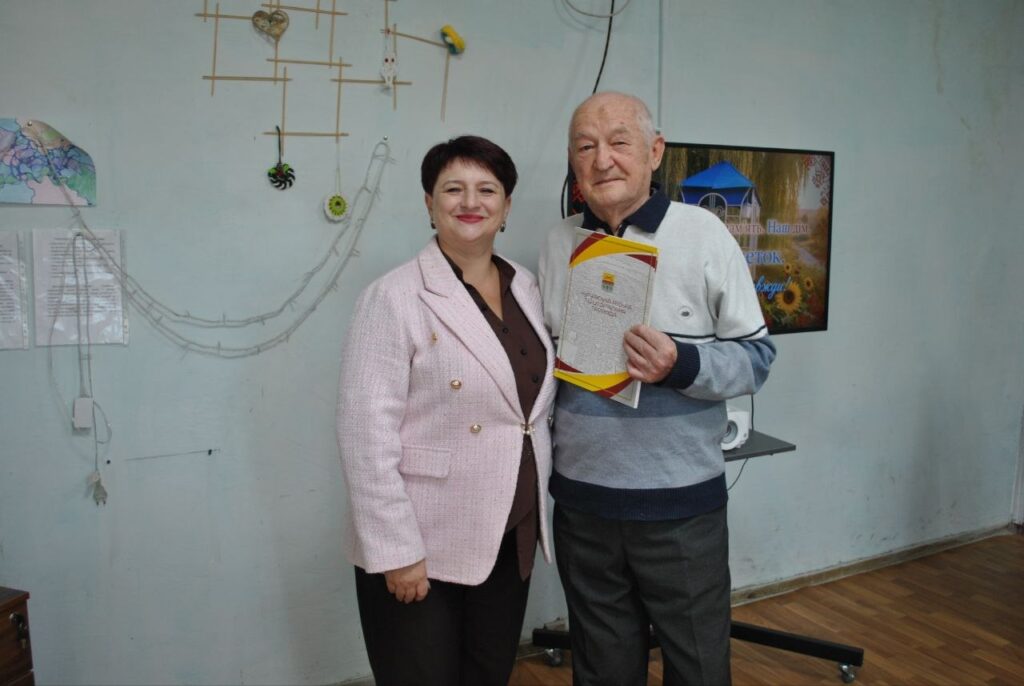 Жінка в рожевому піджаку та старший чоловік у светрі тримають сертифікат на тлі стіни з декором.
