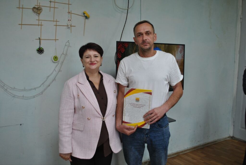 Чоловік у білій футболці тримає сертифікат, стоячи поруч із жінкою в рожевому жакеті.