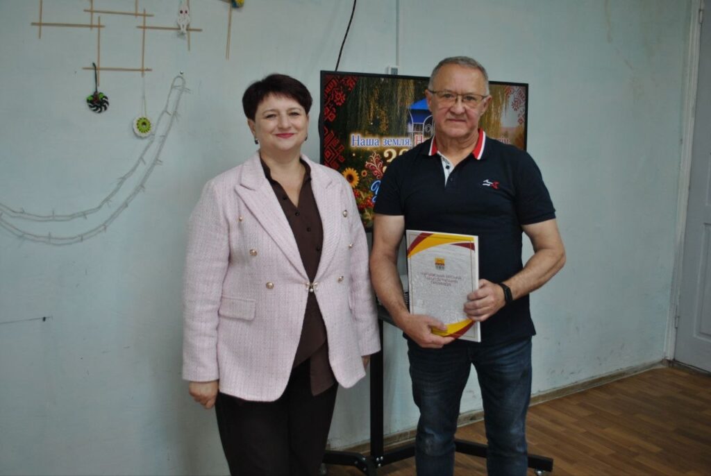 Чоловік у чорній футболці тримає сертифікат, стоячи поруч із жінкою в рожевому піджаку на тлі експозиції з написом «Наша земля».