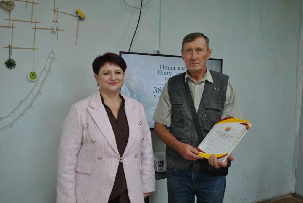 Чоловік у жилеті тримає нагородний документ, стоячи поруч із жінкою в рожевому пальті на тлі екрана з написом «Наша земля».