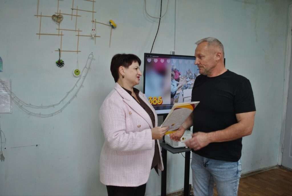 Жінка в рожевому піджаку передає чоловіку в чорній футболці жовту папку з документами.
