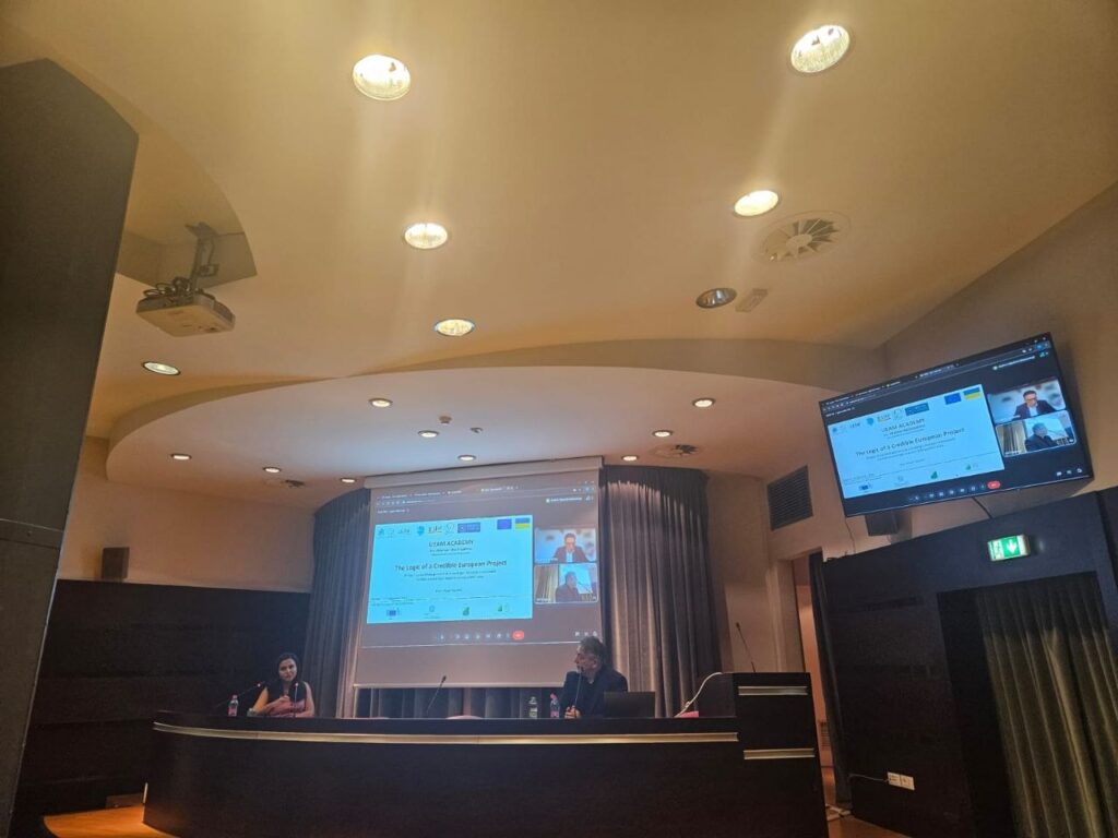 Учасники гібридної конференції «Ukrainian Academy of a Creative European Project» працюють за пультом, дивлячись на екрани з відеотрансляцією.