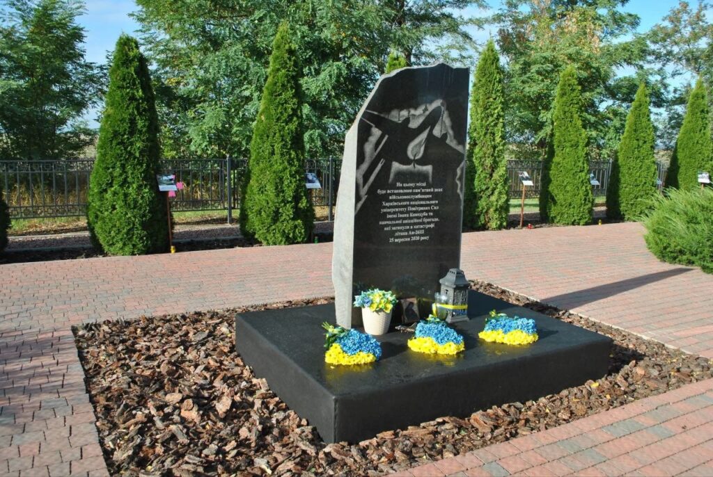 Меморіальний комплекс з чорним обеліском, на якому зображено літак та напис про загиблих у катастрофі Ан-26, прикрашений синьо-жовтими квітами та свічками.