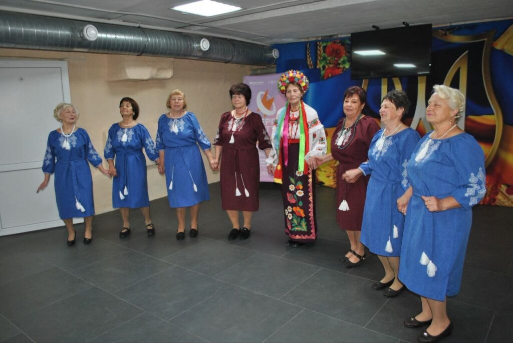 Група жінок у традиційних синіх та бордових сукнях з вишивкою, а також одна жінка в яскравому національному вбранні з вінком, стоять у колі, тримаючись за руки.
