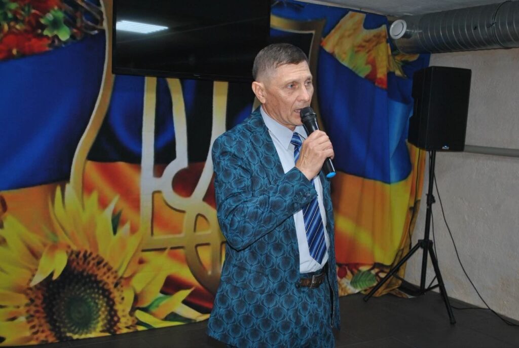 Чоловік у синьому костюмі виступає з мікрофоном на тлі банера з соняшниками та гербом України.