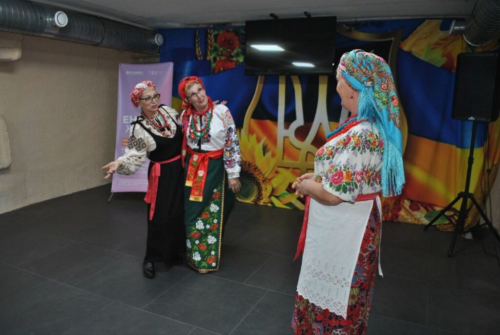 Жінки в традиційних українських вишитих сорочках та головних уборах спілкуються на заході перед банером з гербом України.