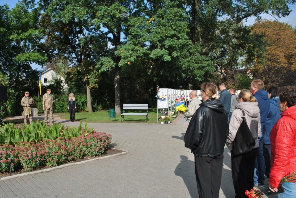 Група людей стоїть у парку біля меморіальної стіни з фото та квітами, де також присутні військові.