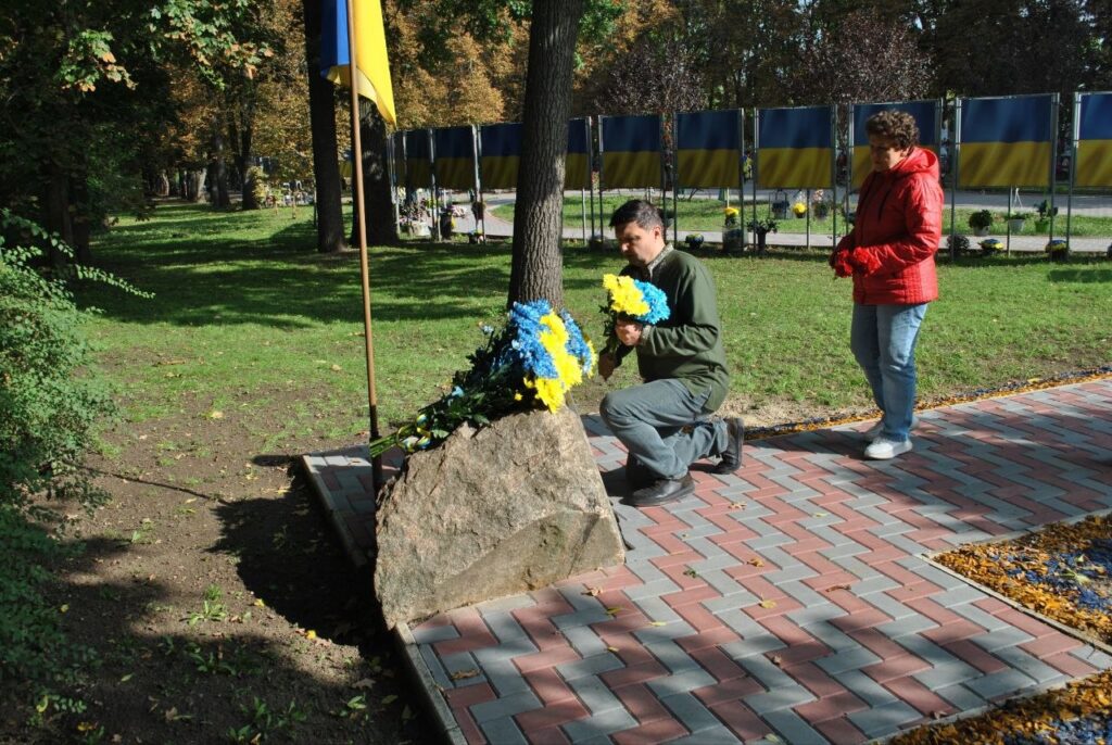 Людина в зеленому светрі стоїть на коліні біля каменю з квітами синьо-жовтого кольору, тримаючи букет, на тлі парку з українськими прапорами.