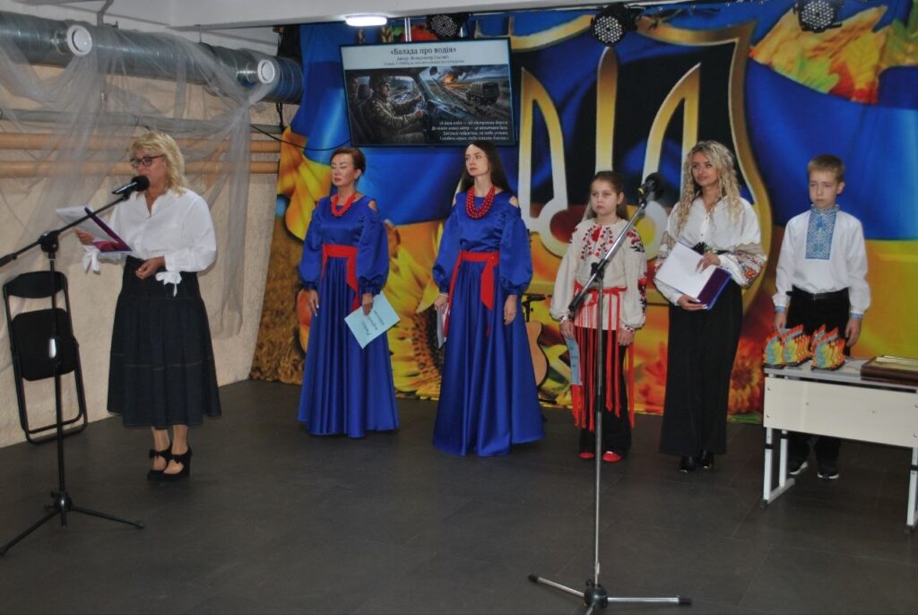 Жінка в білій сорочці читає текст у мікрофон, стоячи на сцені поруч із групою дітей у синіх та білих національних костюмах, на тлі екрана з назвою «Балада про подія».