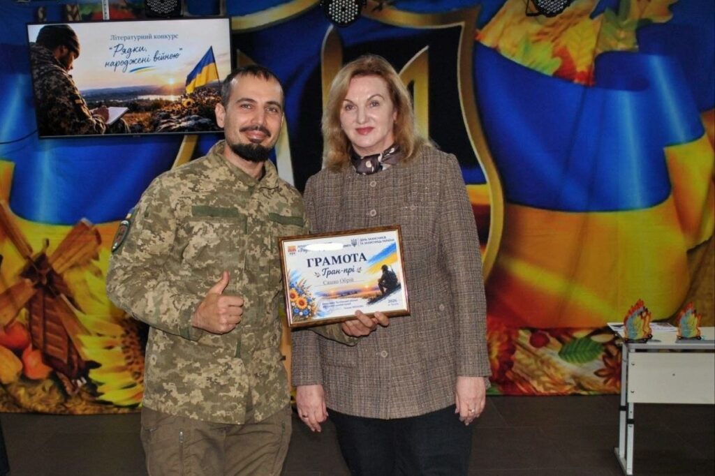 Військовослужбовець у камуфляжі та жінка в пальті тримають нагороду з написом «ГРАМОТА Гран-прі» на тлі банера літературного конкурсу «Рядки, народжені війною».
