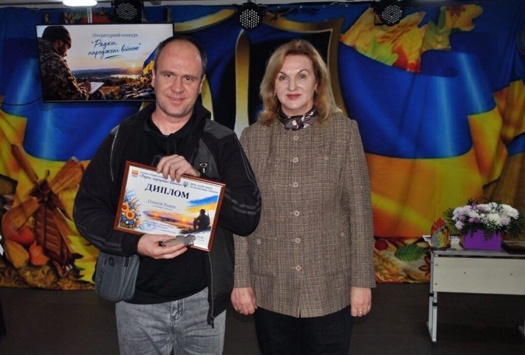 Чоловік тримає диплом літературного конкурсу «Родки, народжені війною» поруч із жінкою на тлі сцени.