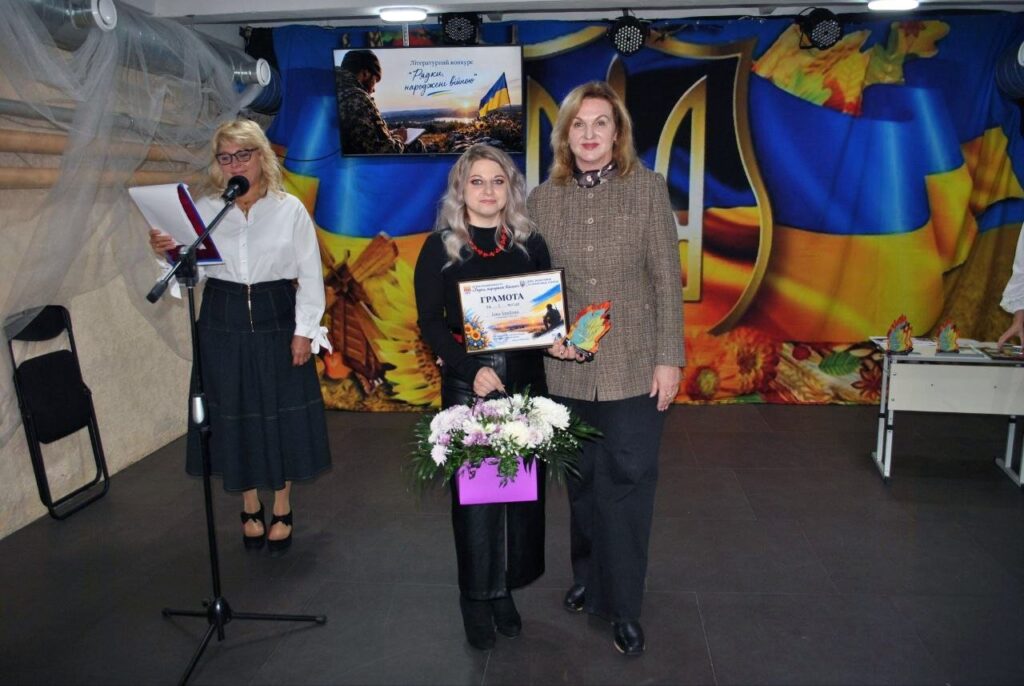 Жінка тримає грамоту та букет квітів, стоячи поруч з іншою жінкою на тлі банера літературного конкурсу «Радки народжених бійцям».