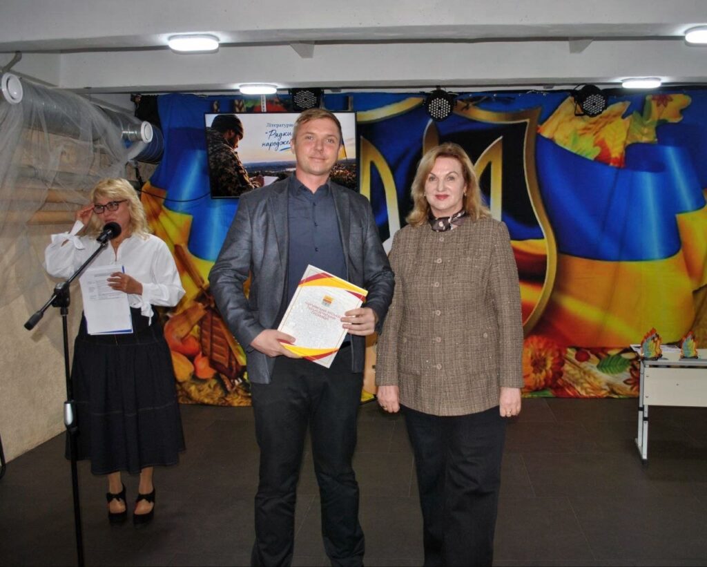 Чоловік тримає диплом, стоячи поруч із жінкою на тлі банера з гербом України, тоді як інша жінка читає текст у мікрофон.