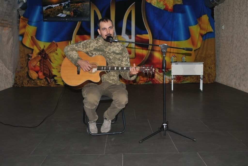 Чоловік у камуфляжі грає на гітарі та співає в мікрофон на тлі українського прапора.