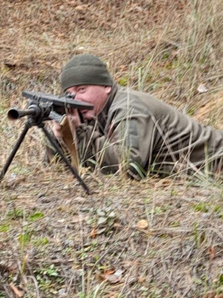 Чоловік у військовому одязі та шапці лежить у траві, прицілюючись зі стрілецької зброї на штативі.